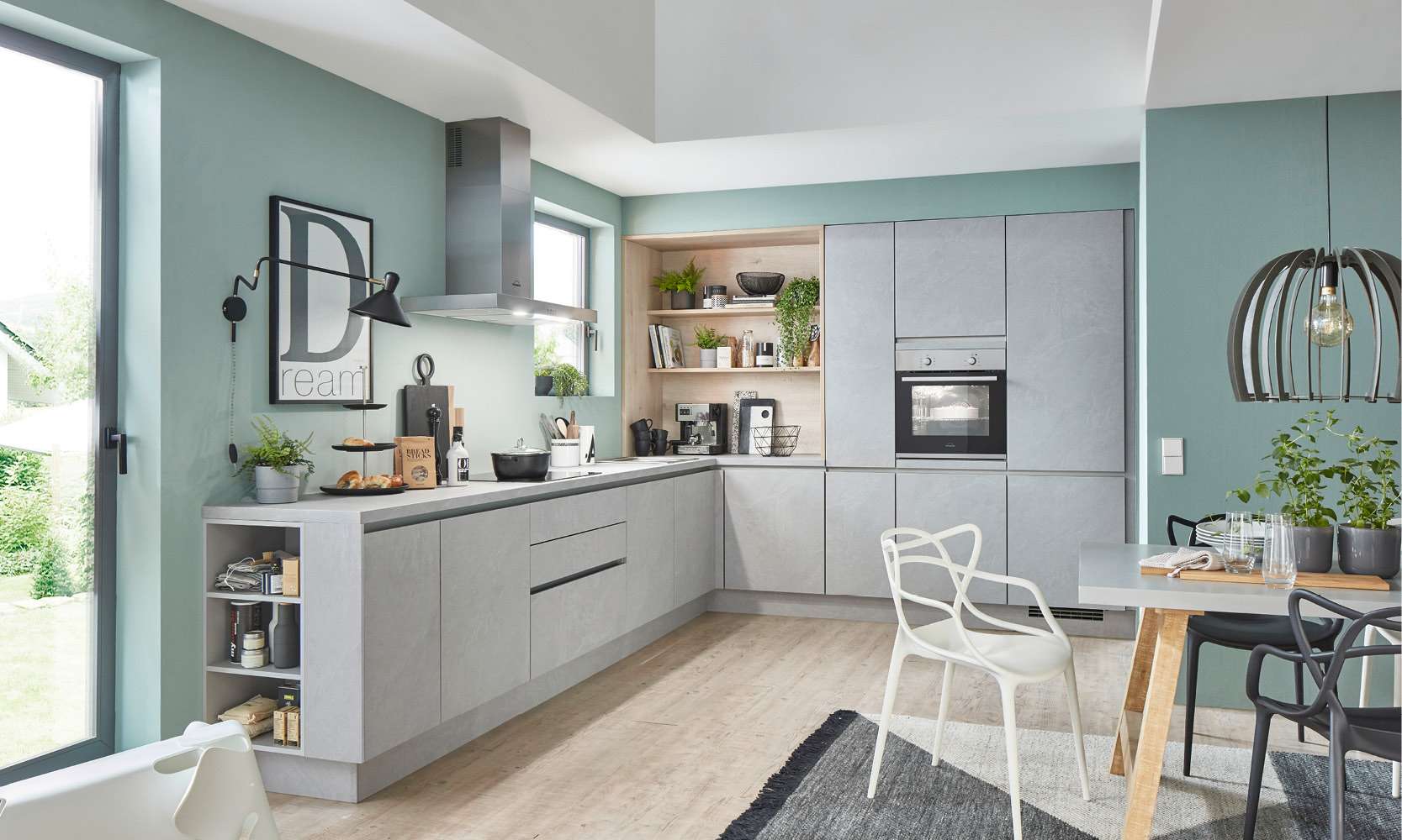 Moderne L-Küche mit matten, hellgrauen Fronten und offener Regalecke aus hellem Holz. Die Küche ist stilvoll eingerichtet mit Dekorationen, Pflanzen und hochwertigen Küchengeräten. Im Essbereich rechts steht ein grauer Tisch mit Designerstühlen vor einer Wand in sanftem Grün. Große Fenster bringen Tageslicht in den Raum.