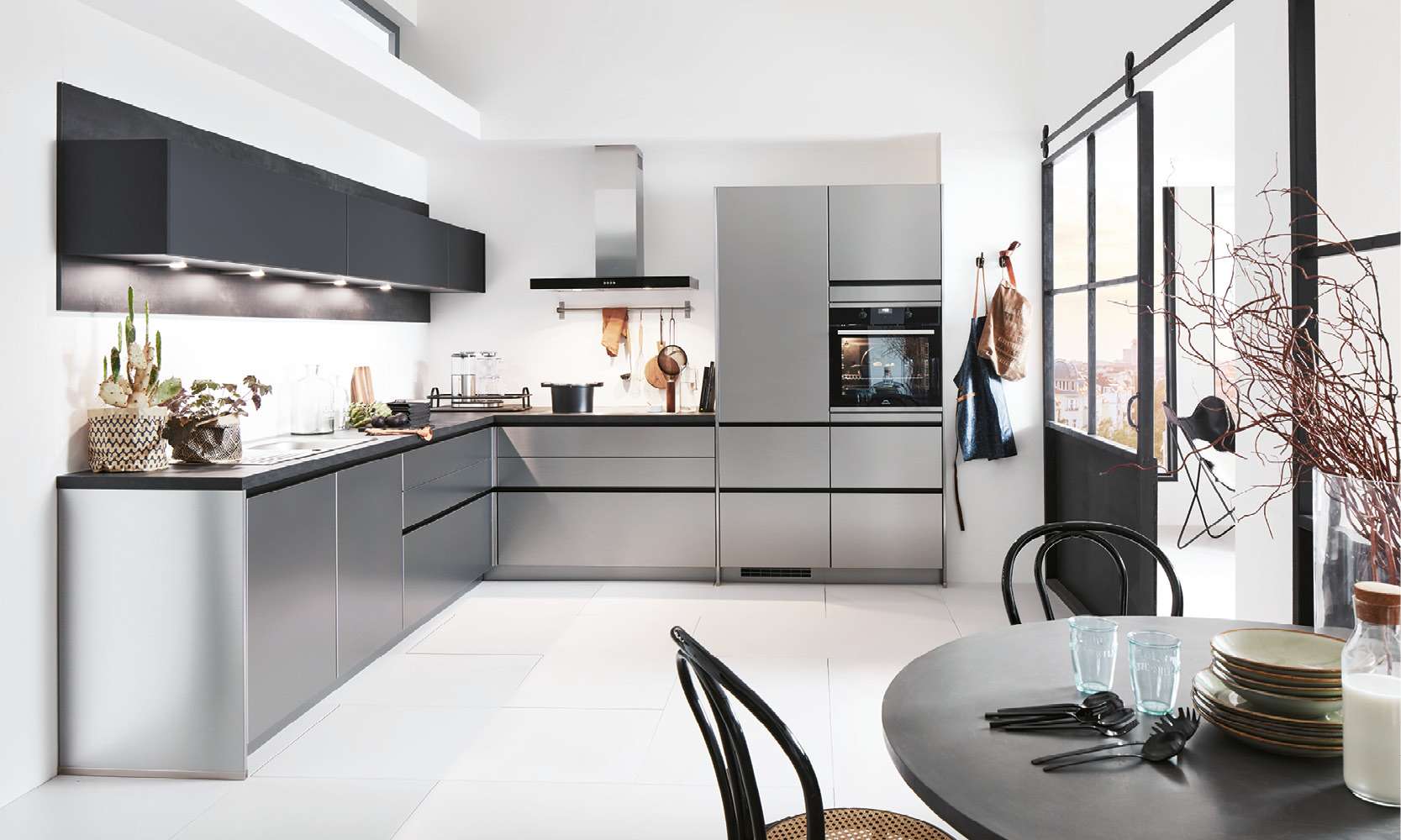 Moderne L-Küche mit grifflosen Fronten in elegantem Grau-Metallic und mattschwarzen Oberschränken. Die Küche ist ausgestattet mit Einbaugeräten, Dunstabzugshaube und stilvollen Accessoires. Im Vordergrund ein gedeckter runder Tisch, im Hintergrund ein schwarzes Schiebetür-Element mit Glas und Ausblick auf die Stadt.