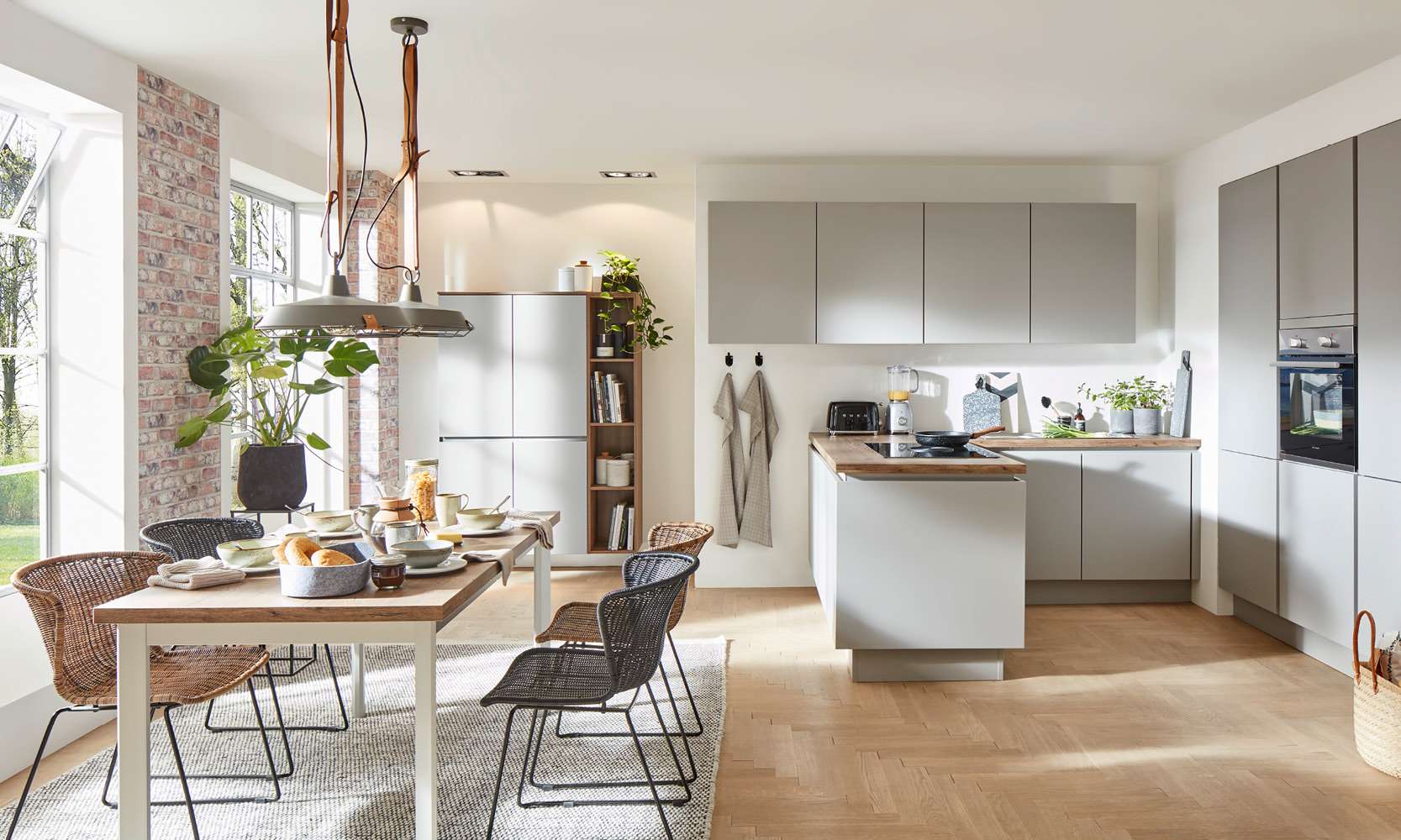 Moderne, helle Einbauküche mit Kücheninsel, grifflosen Fronten in Mattgrau und Holzarbeitsplatte. Links im Bild ein gedeckter Esstisch mit vier Stühlen, großen Fenstern und Ziegelwand. Die Küche ist wohnlich dekoriert mit Pflanzen, Kochutensilien und einem offenen Regal.