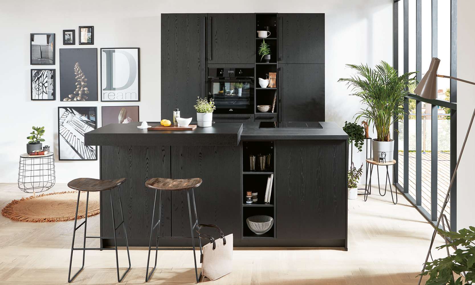 Moderne, stilvolle Designküche in elegantem Schwarz mit Kücheninsel, integriertem Backofen und offenem Regal. Die Küche ist minimalistisch eingerichtet, mit grünen Pflanzen, Barhockern aus Holz und Metall sowie großem Lichteinfall durch bodentiefe Fenster. Im Hintergrund eine dekorative Bilderwand.