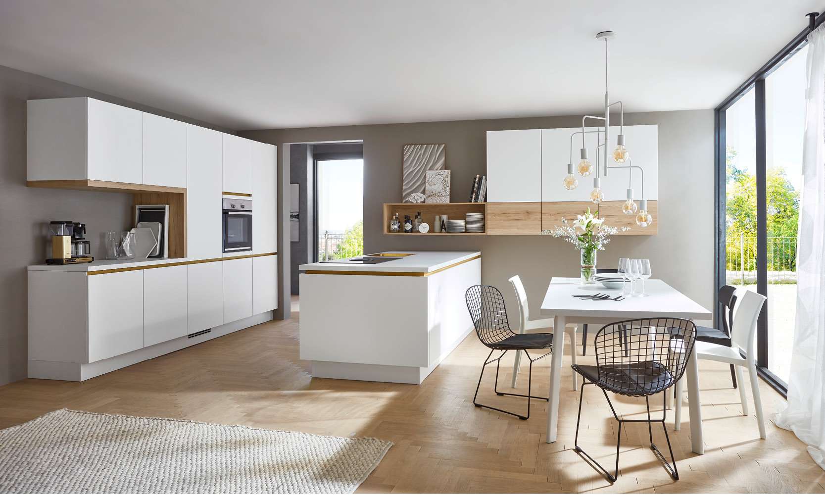 Helle Designküche mit weißen, grifflosen Fronten und warmen Holzelementen. Die Küche verfügt über eine moderne Insel mit Kochfeld, offene Regale und einen angrenzenden Essbereich mit weißem Tisch und schwarzen Drahtstühlen. Große Fenster sorgen für viel Tageslicht und Blick ins Grüne.