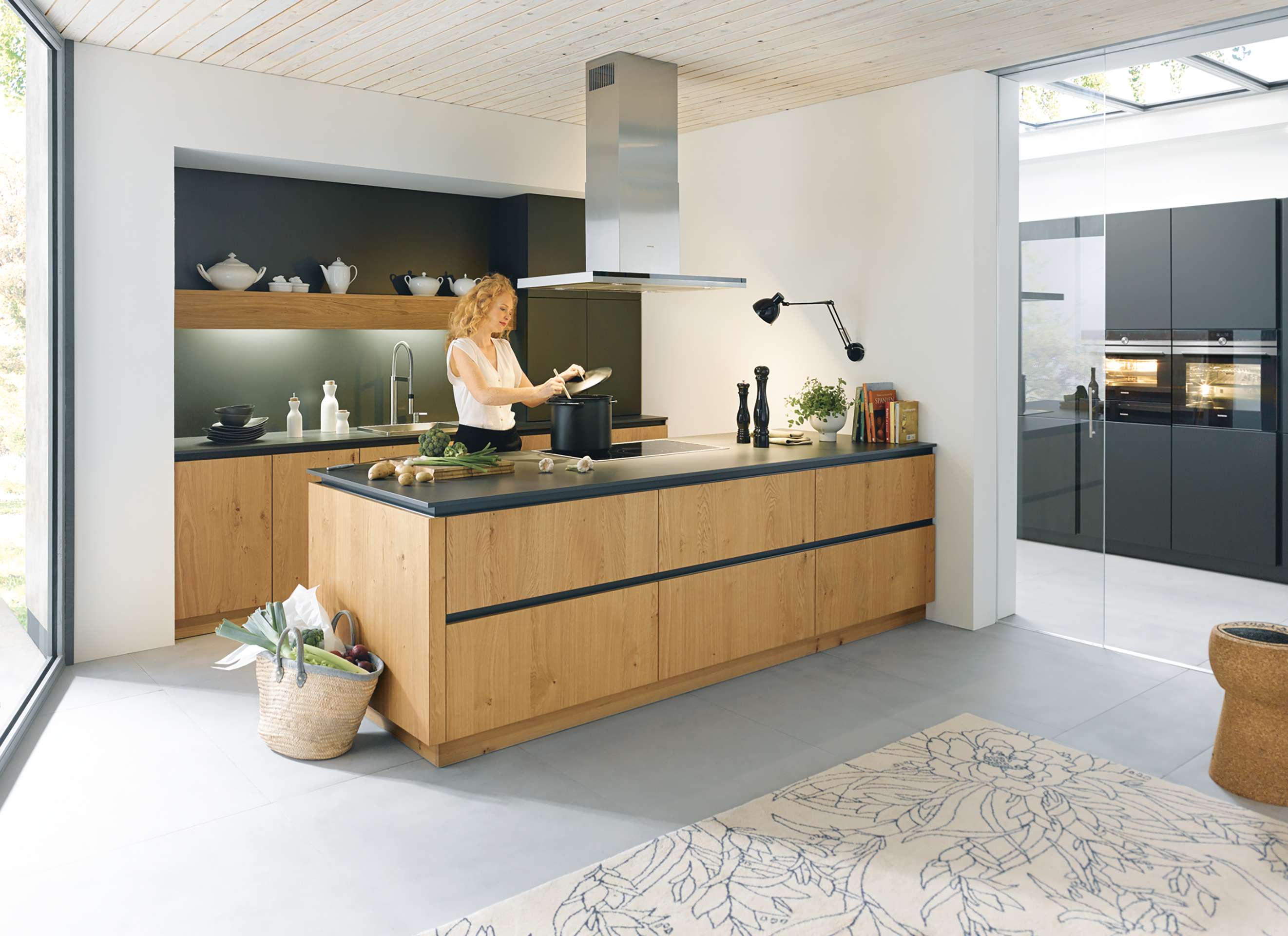 Schüller Küche mit Holzinsel, dunklen Akzenten und großer Fensterfront, in der eine Frau kocht.
