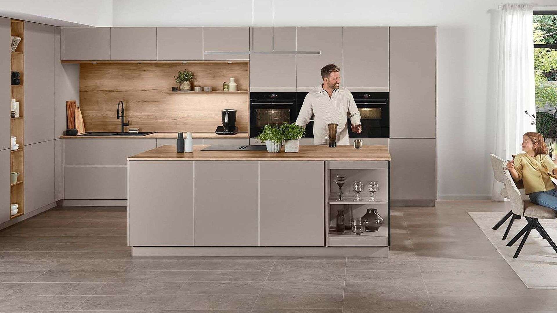 Moderne beige Wohnküche mit Holzakzenten, Kochinsel mit integriertem Glasregal, Mann beim Kochen, Frau am angrenzenden Esstisch, große Fensterfronten mit Gartenblick.