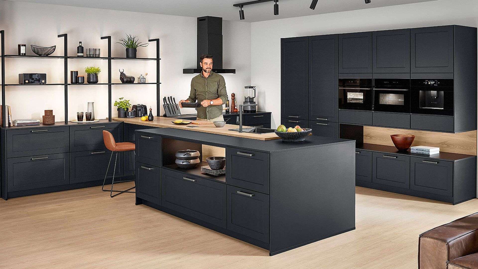 Moderne anthrazitfarbene Küche mit Kochinsel und Holzarbeitsplatten, Mann am Kochen, offene Metallregale mit beleuchteten Böden links, Hochschränke mit integrierten Einbaugeräten rechts, Arbeitsplatz mit ockerfarbenem Stuhl.