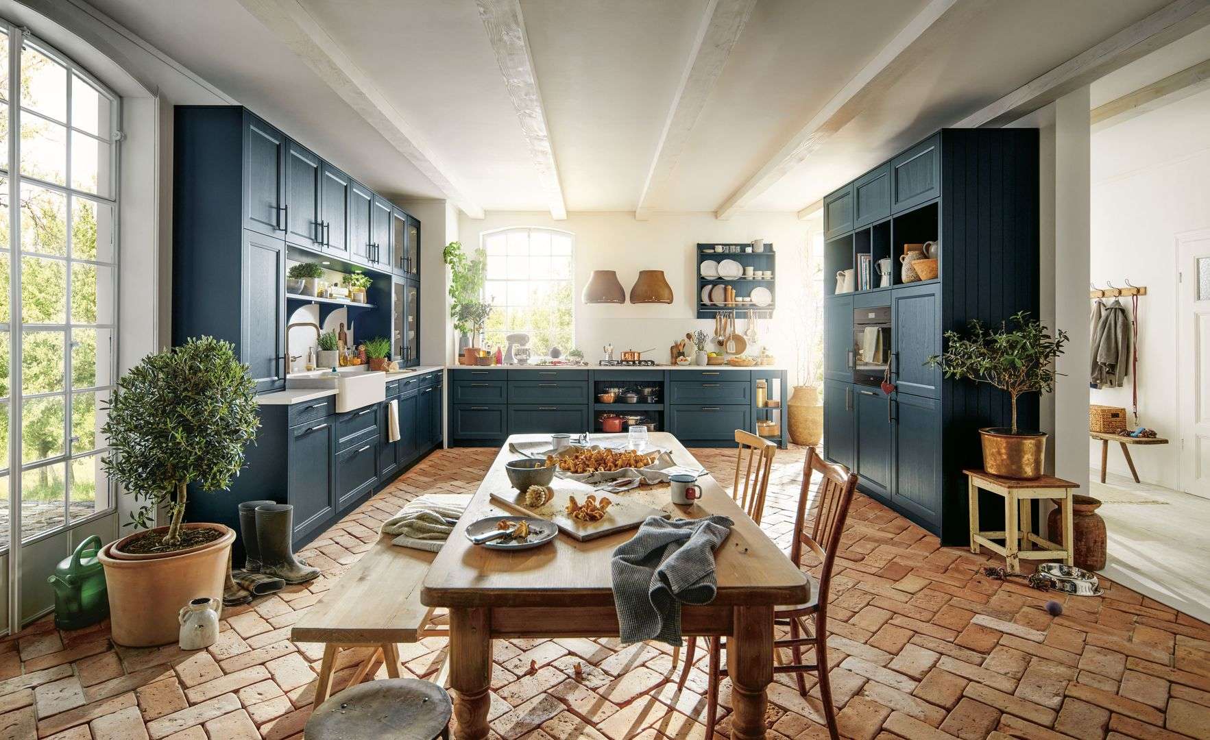 Landhausküche von Schüller mit dunkelblauen Küchenschränken, großem Holztisch in der Mitte, Terrakottaboden und großen Fenstern mit Blick ins Grüne.