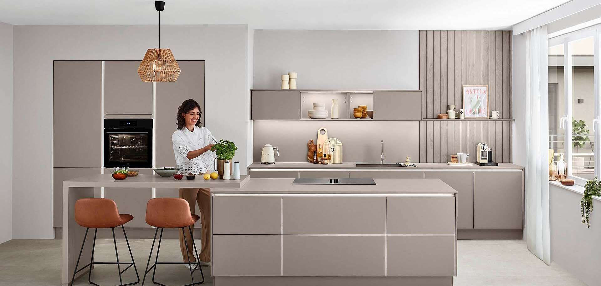 Moderne beige Küche mit Kochinsel, Frau in weißem Kleid beim Kochen mit Kräutern, zwei rostfarbene Barhocker, Korbpendelleuchte, Hochschränke mit Backofen links, beleuchtete Hängeschränke mit Glastüren und Durchreiche zum Nebenraum
