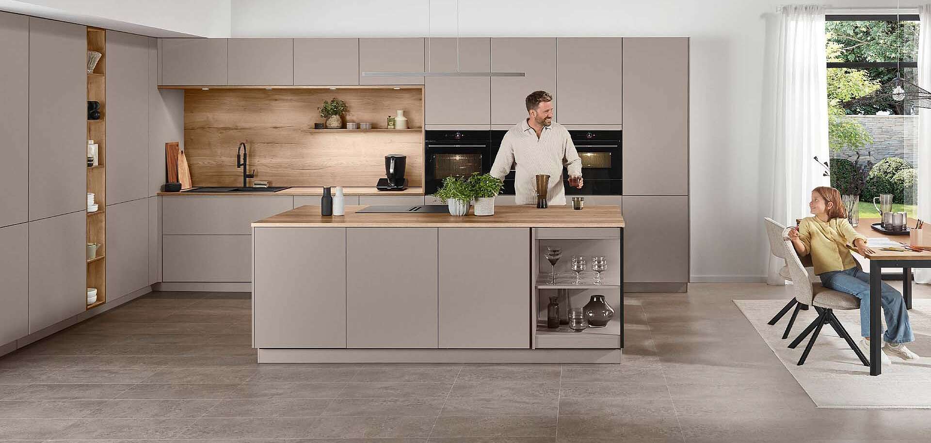 Moderne beige Wohnküche mit Holzakzenten, Kochinsel mit integriertem Glasregal, Mann beim Kochen, Frau am angrenzenden Esstisch, große Fensterfronten mit Gartenblick.