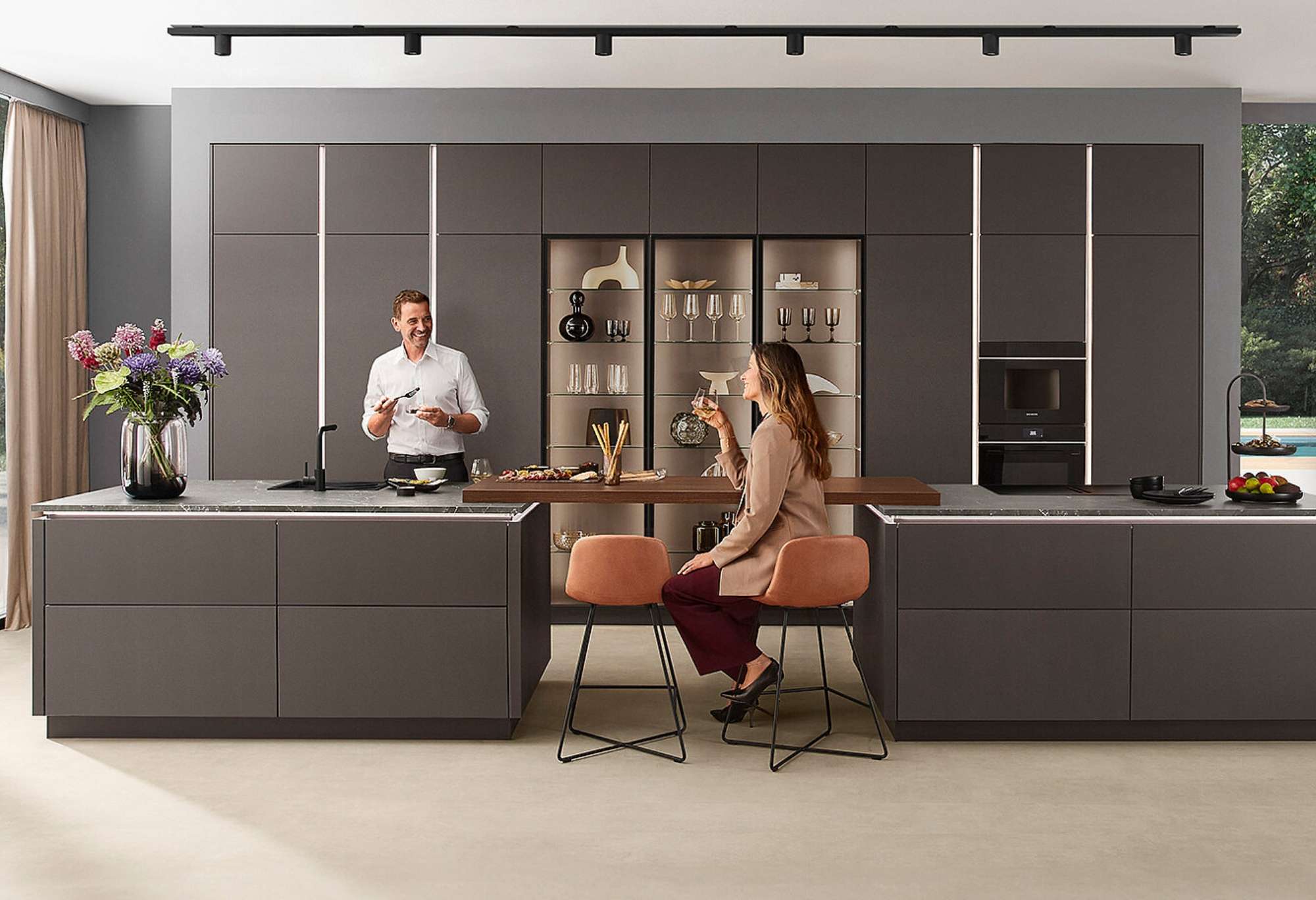 Moderne anthrazitfarbene Designerküche mit zwei großen Kücheninseln, beleuchteten Vitrinen-Oberschränken, integrierten Einbaugeräten, Paar im Gespräch an rostfarbenen Barhockern, großzügige Glasfronten mit Gartenblick beidseitig