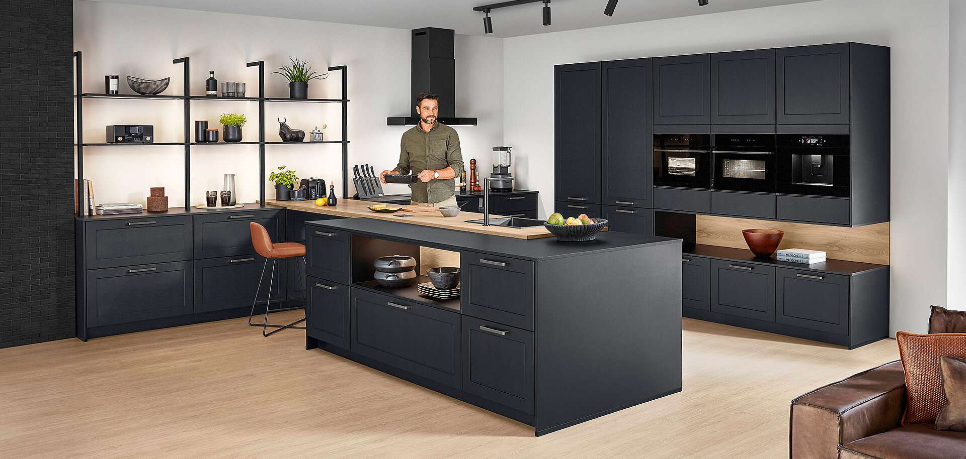 Moderne anthrazitfarbene Küche mit Kochinsel und Holzarbeitsplatten, Mann am Kochen, offene Metallregale mit beleuchteten Böden links, Hochschränke mit integrierten Einbaugeräten rechts, Arbeitsplatz mit ockerfarbenem Stuhl.