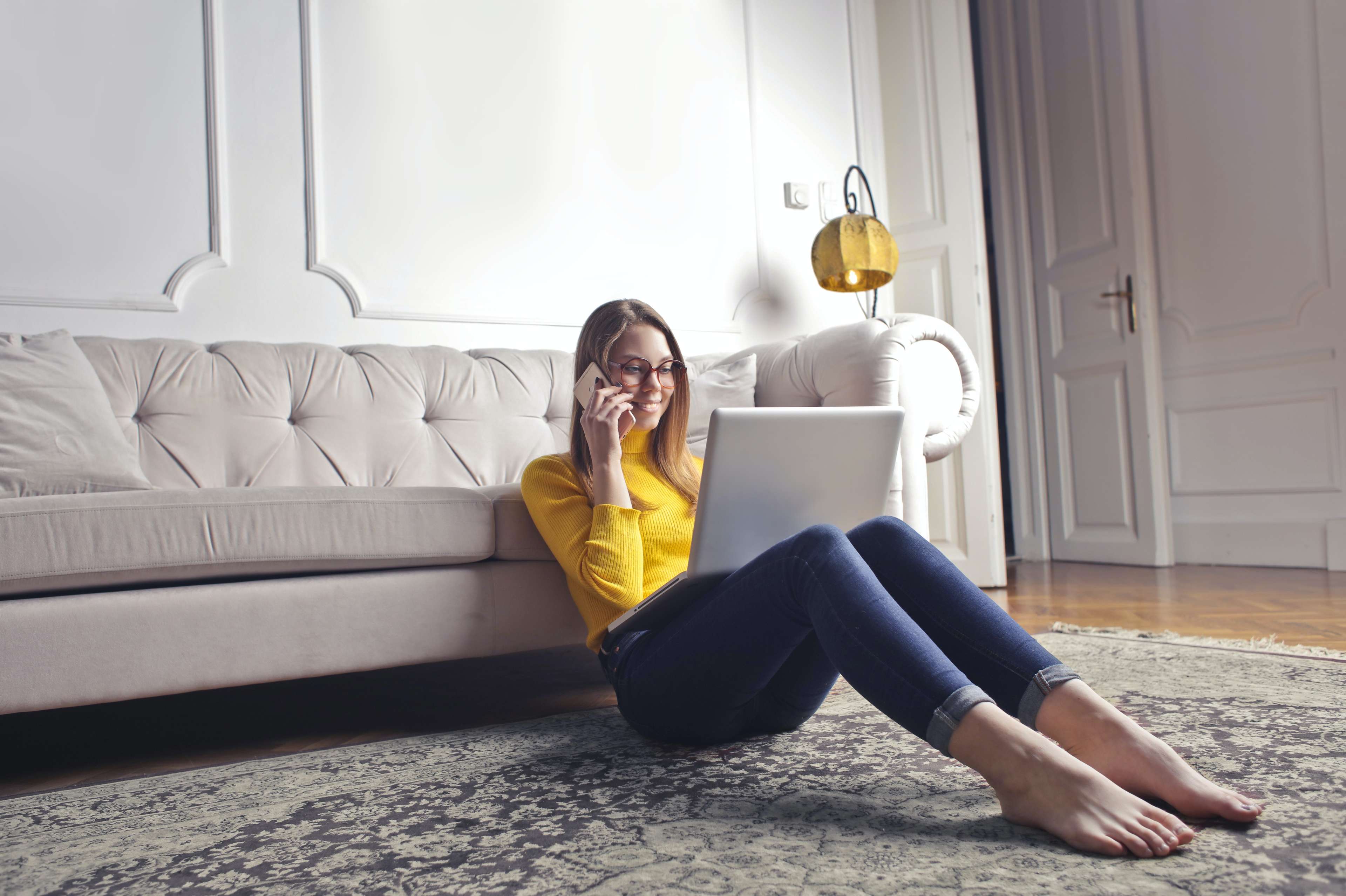 Junge Frau am Laptop sitzt telefonierend vor einer weißen Couch
